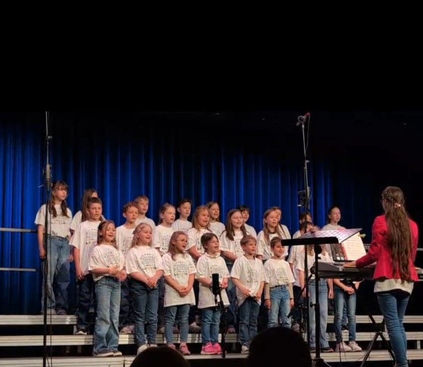 Ein Chor junger Mädchen und Jungen tritt auf der Bühne auf, begleitet von einer Frau am Klavier. Sie tragen passende weiße Shirts und blaue Jeans. Die Bühne ist mit blauen Vorhängen geschmückt.