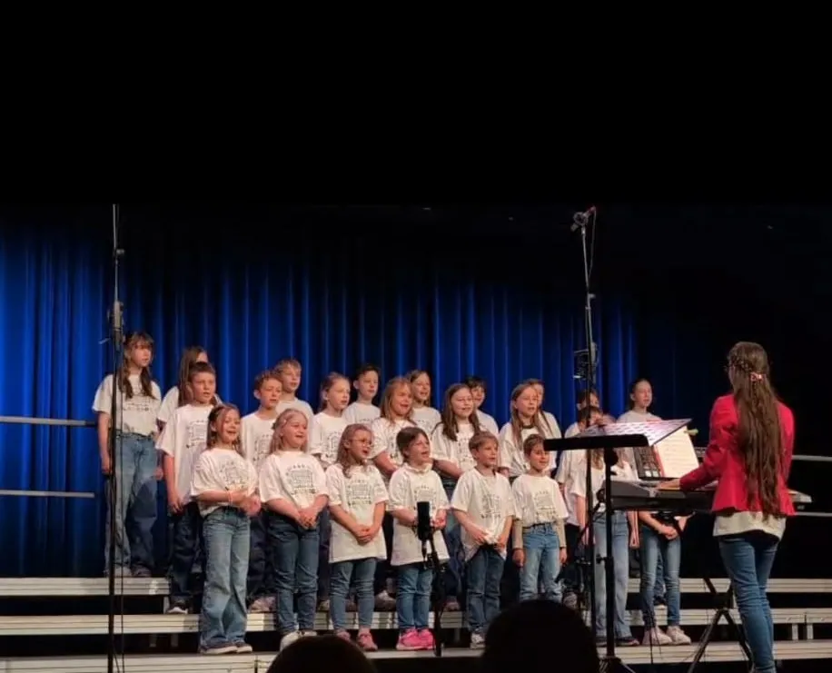 Ein Chor junger Mädchen und Jungen tritt auf der Bühne auf, begleitet von einer Frau am Klavier. Sie tragen passende weiße Shirts und blaue Jeans. Die Bühne ist mit blauen Vorhängen geschmückt.