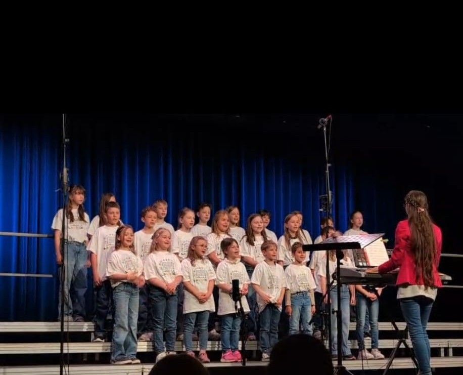 Ein Chor junger Mädchen und Jungen tritt auf der Bühne auf, begleitet von einer Frau am Klavier. Sie tragen passende weiße Shirts und blaue Jeans. Die Bühne ist mit blauen Vorhängen geschmückt.