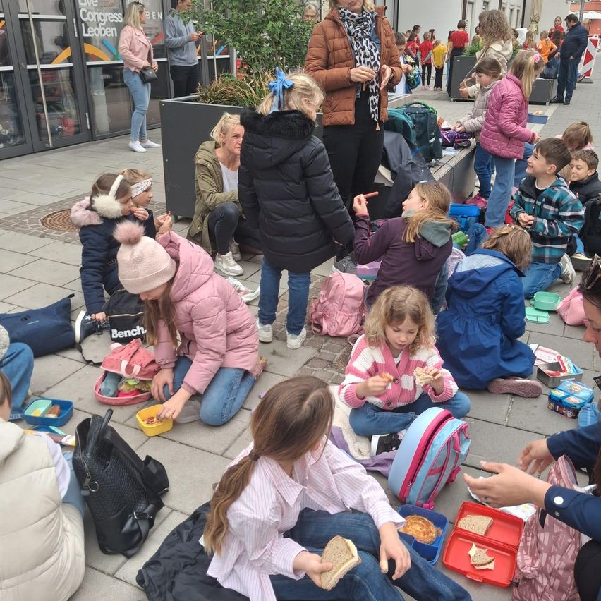 Eine Gruppe von Kindern und Erwachsenen sitzt am Boden und isst. Eine Frau steht und spricht mit einem Kind. Ein Topf mit einer Pflanze steht auf dem Bürgersteig. In der Umgebung befinden sich mehrere Gebäude.