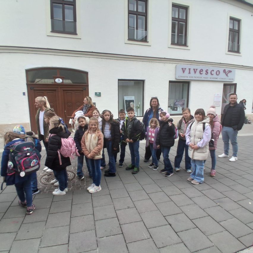 Eine Gruppe von Kindern und Erwachsenen steht vor einem Gebäude mit einem Schild Vivesco. Die Kinder tragen Jacken und Rucksäcke. Einige von ihnen halten Händchen.