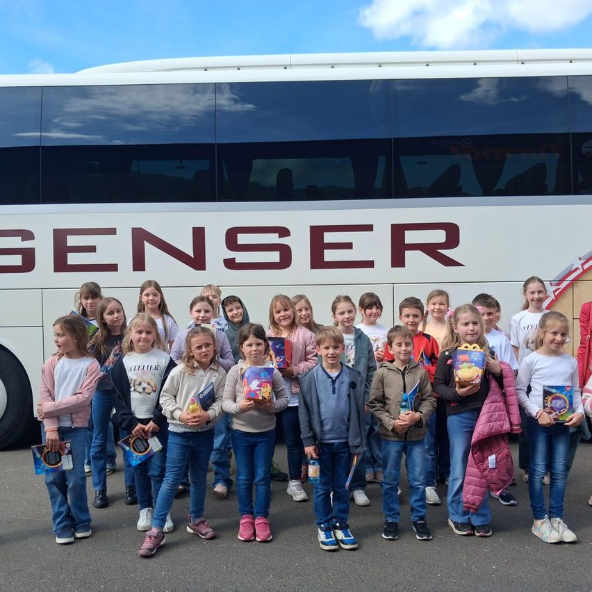 Eine Gruppe von Kindern posiert für ein Foto vor einem weißen Bus mit roten Buchstaben, die 'GENSER' lesen. Jedes Kind hält ein Buch oder eine McDonald's-Tasche.