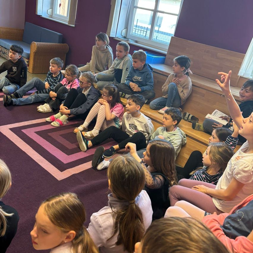 Eine Gruppe von Kindern sitzt auf Holzbänken in einem Raum, wobei ein Mädchen die Hand hebt und andere zuschauen. Es gibt einen gemusterten Teppich auf dem Boden.