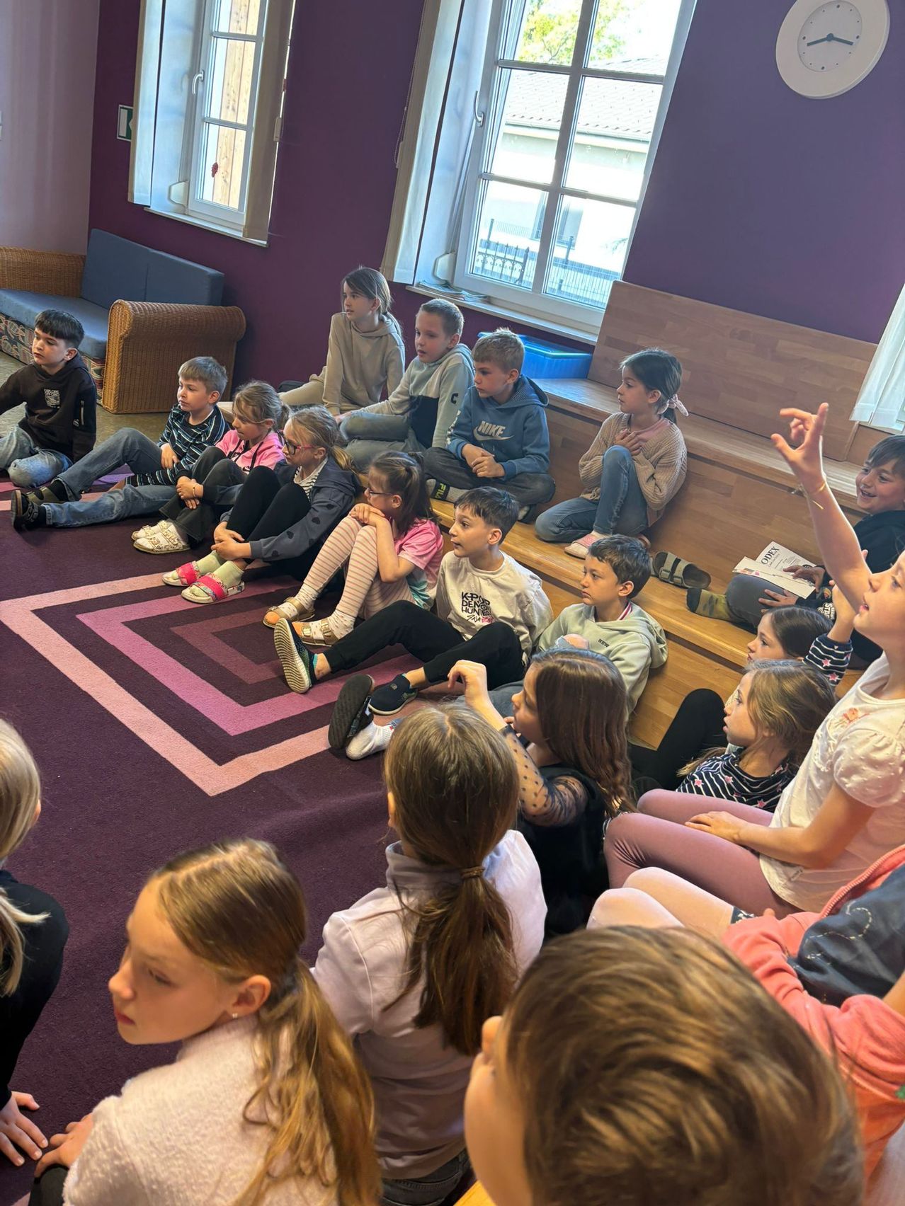 Eine Gruppe von Kindern sitzt auf Holzbänken in einem Raum, wobei ein Mädchen die Hand hebt und andere zuschauen. Es gibt einen gemusterten Teppich auf dem Boden.