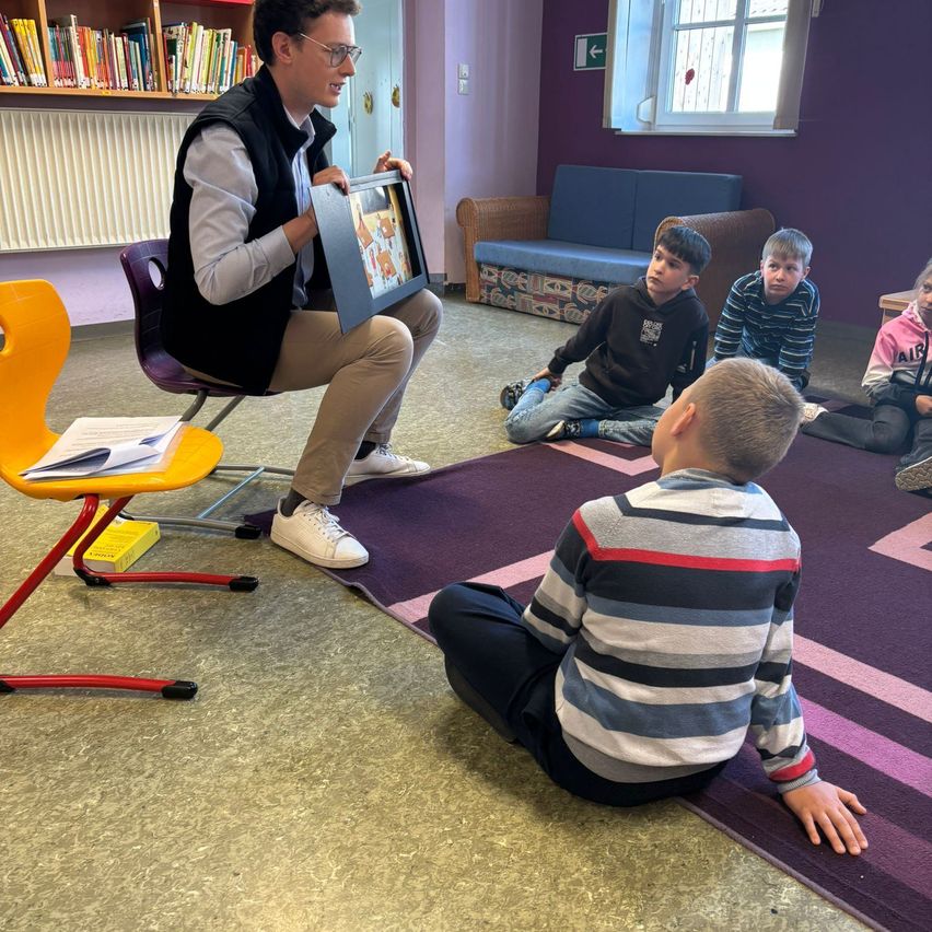 Ein Mann hält ein gerahmtes Bild vor eine Gruppe von Kindern, die auf dem Boden sitzen.