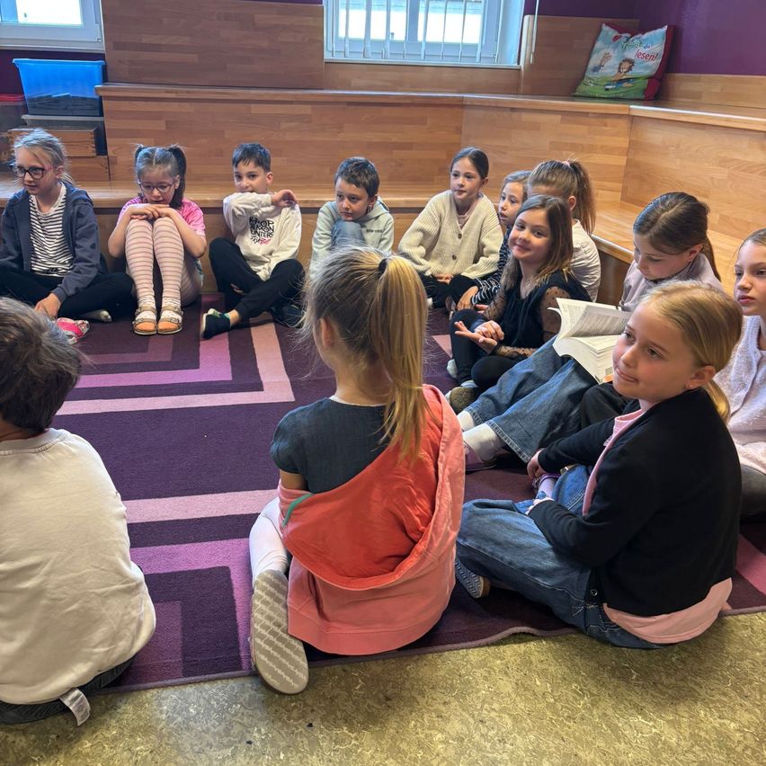 Eine Gruppe von Kindern sitzt im Kreis auf dem Boden, einige lesen Bücher. Ein Mädchen mit einer pinken Tasche schaut in ein Buch.