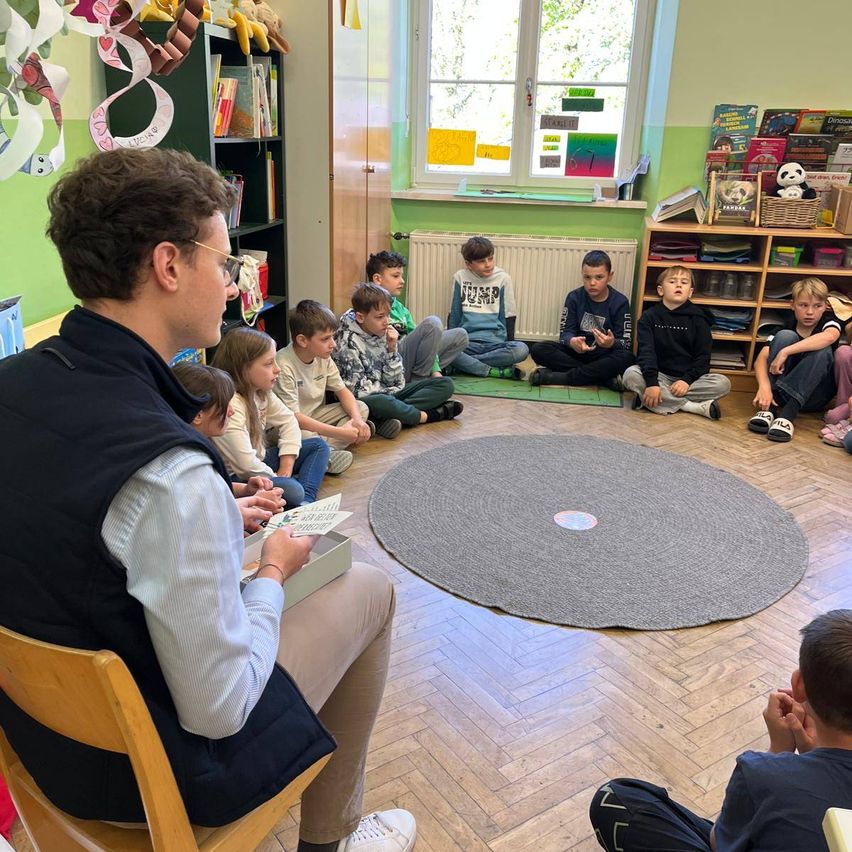 Ein Mann liest ein Buch vor einer Gruppe von Kindern, die auf dem Boden in einem Klassenzimmer sitzen. Die Kinder sind aufmerksam. Ein großer runder Teppich liegt auf dem Boden.