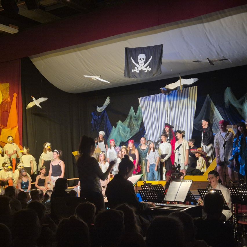 Eine Aufführung auf einer Bühne mit einer Piratenflagge. Eine Gruppe von Kindern in Piratenkostümen tritt vor einem Publikum auf. Eine Frau steht vor einem Klavier, und ein Mann spielt Saxophon.