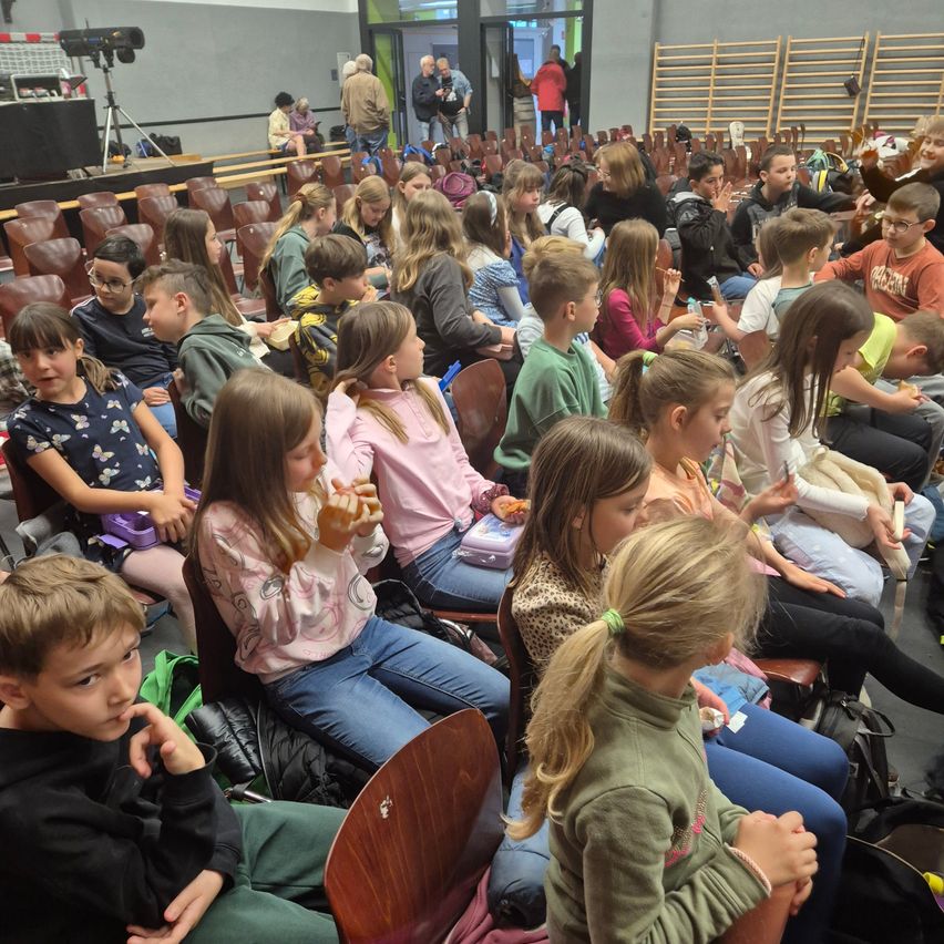 Eine Gruppe von Kindern und Erwachsenen sitzt in einem Auditorium. Einige der Kinder essen Snacks und andere schauen sich etwas an. Der Raum ist gut beleuchtet und hat Holzstühle und eine Bühne mit einer Kamera.
