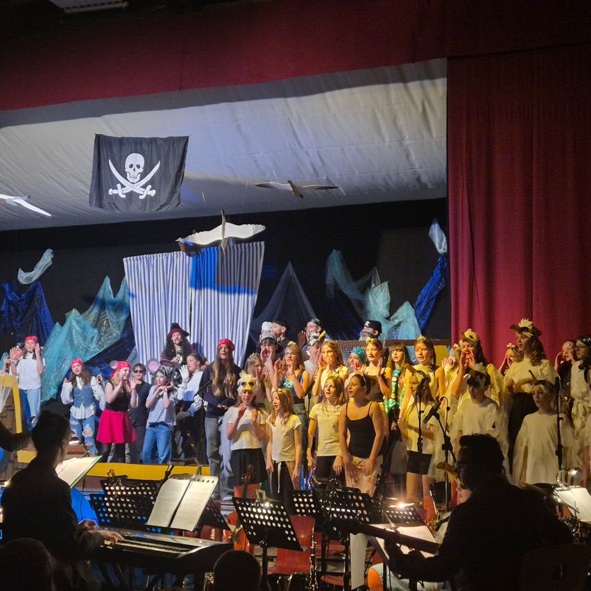 Ein Chor in Piratenkleidung tritt mit Musikinstrumenten auf der Bühne auf, im Hintergrund weht eine Piratenflagge.