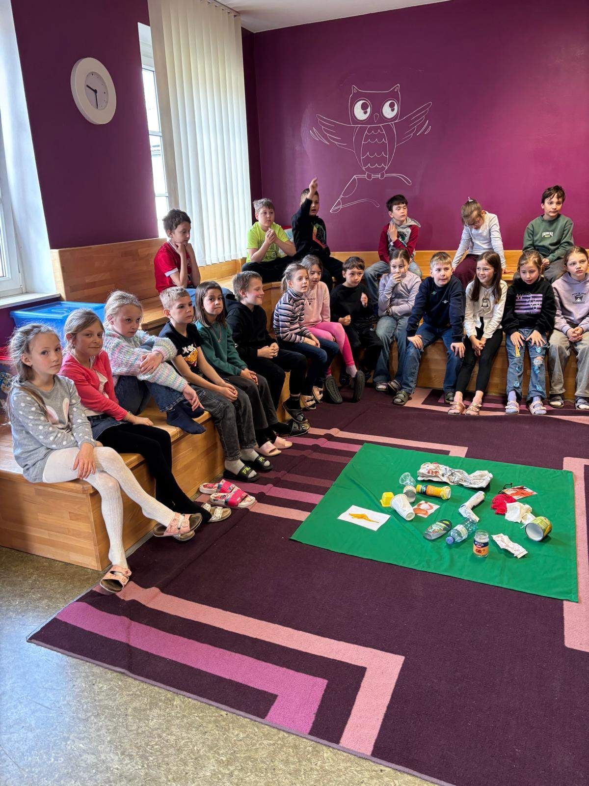 Ein Klassenzimmer gefüllt mit Kindern, die auf Bänken sitzen, mit verschiedenen Abfallgegenständen auf einem grünen Teppich vor ihnen.