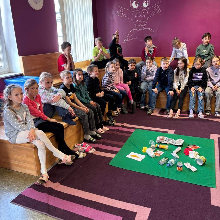 Ein Klassenzimmer gefüllt mit Kindern, die auf Bänken sitzen, mit verschiedenen Abfallgegenständen auf einem grünen Teppich vor ihnen.