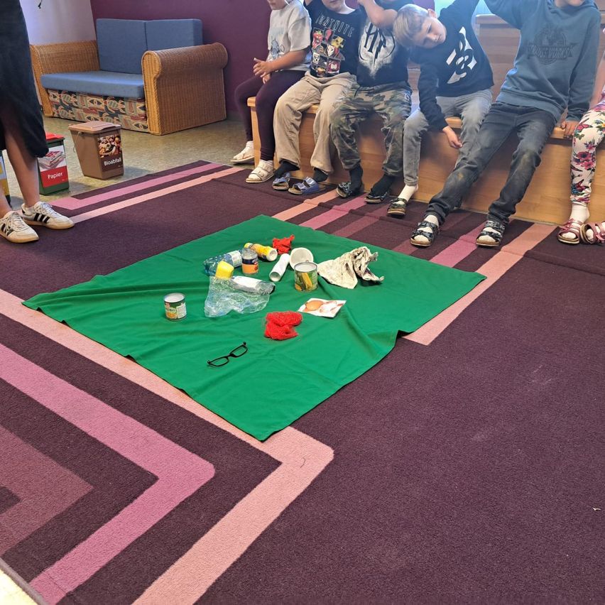 Mehrere Kinder sitzen auf einer Bank in einem Raum mit Teppichboden. Ein grüner Stoff ist auf dem Teppich ausgebreitet, mit verschiedenen Gegenständen darauf, einschließlich einer Dose, einer Plastikflasche, einem Becher und einer Brille. Ein Mülleimer steht in der Nähe.