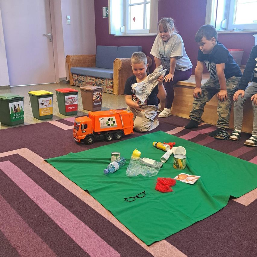 Kinder spielen mit einem Recycling-Lkw auf einer grünen Decke, umgeben von Mülltonnen und Recycling-Artikeln.