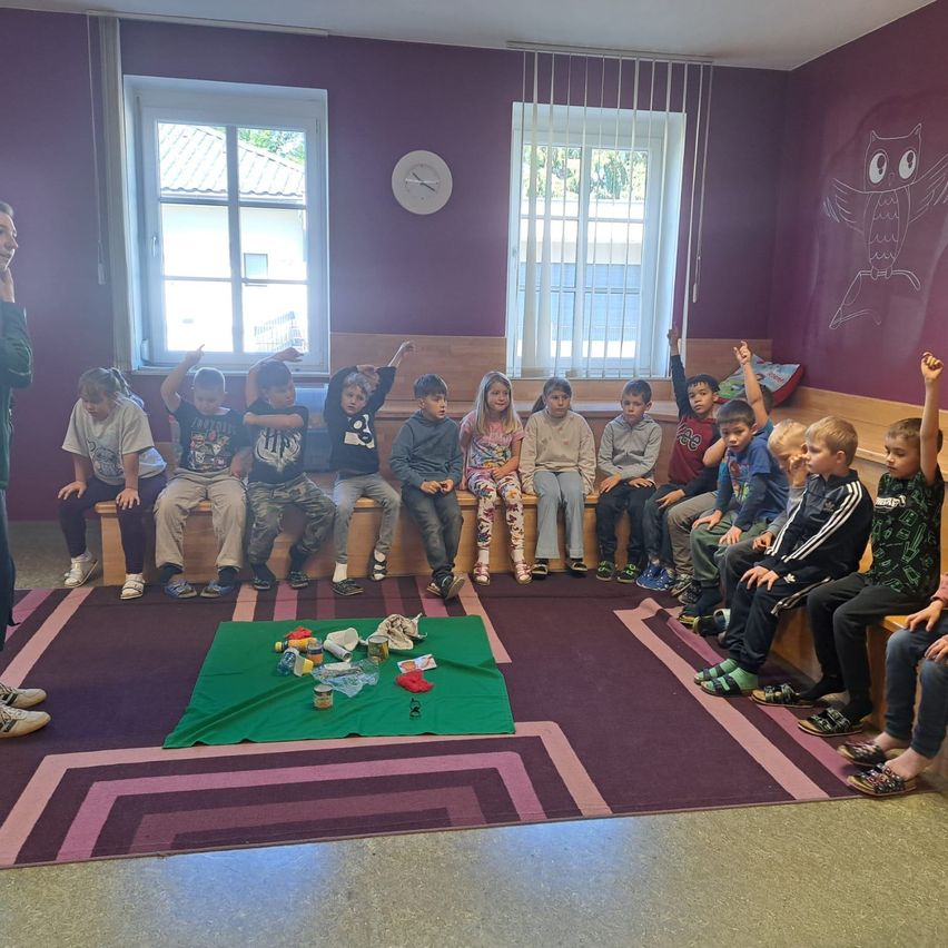 Eine Gruppe von Kindern sitzt auf einem Teppich in einem Kreis, mit Spielzeug und Snacks in der Mitte. Sie sind lässig gekleidet, und ein Kind hebt eine Hand. Der Raum hat lila Wände, eine Uhr und eine Eule-Zeichnung.