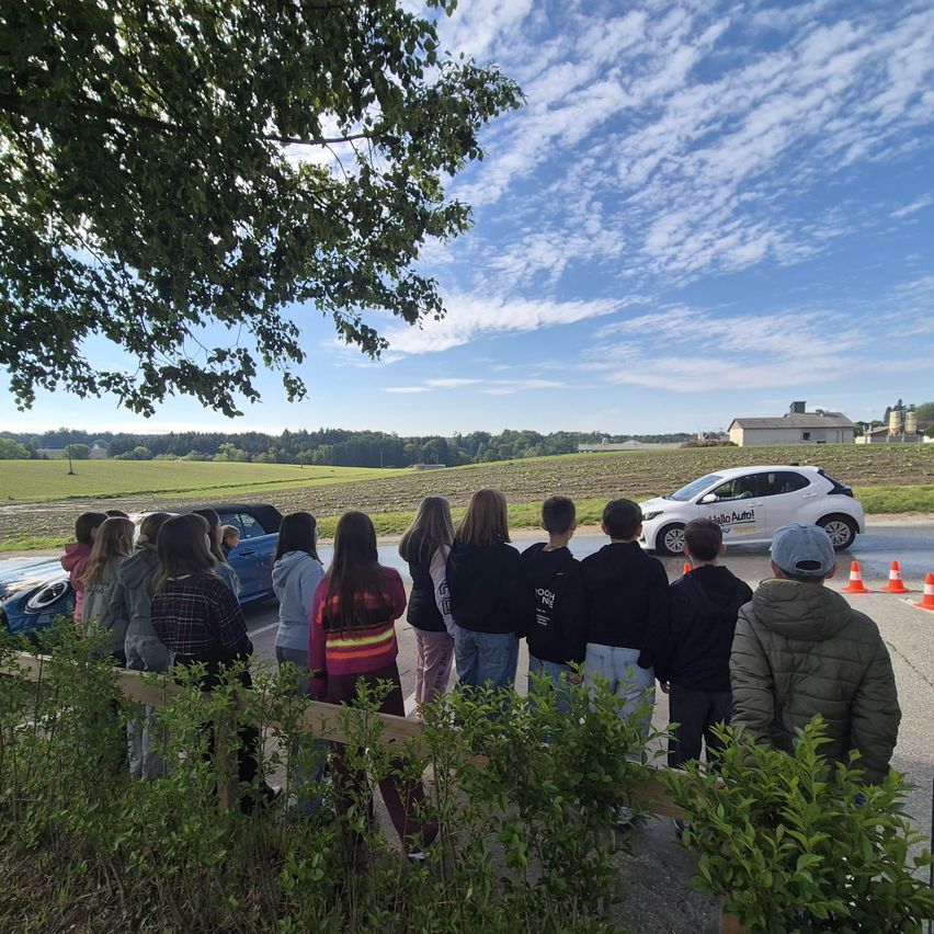 Eine Gruppe junger Menschen steht am Straßenrand und beobachtet, wie ein weißer Wagen vorbeifährt. Das Gebiet ist von Grün umgeben, und in der Ferne ist ein Gebäude zu sehen.