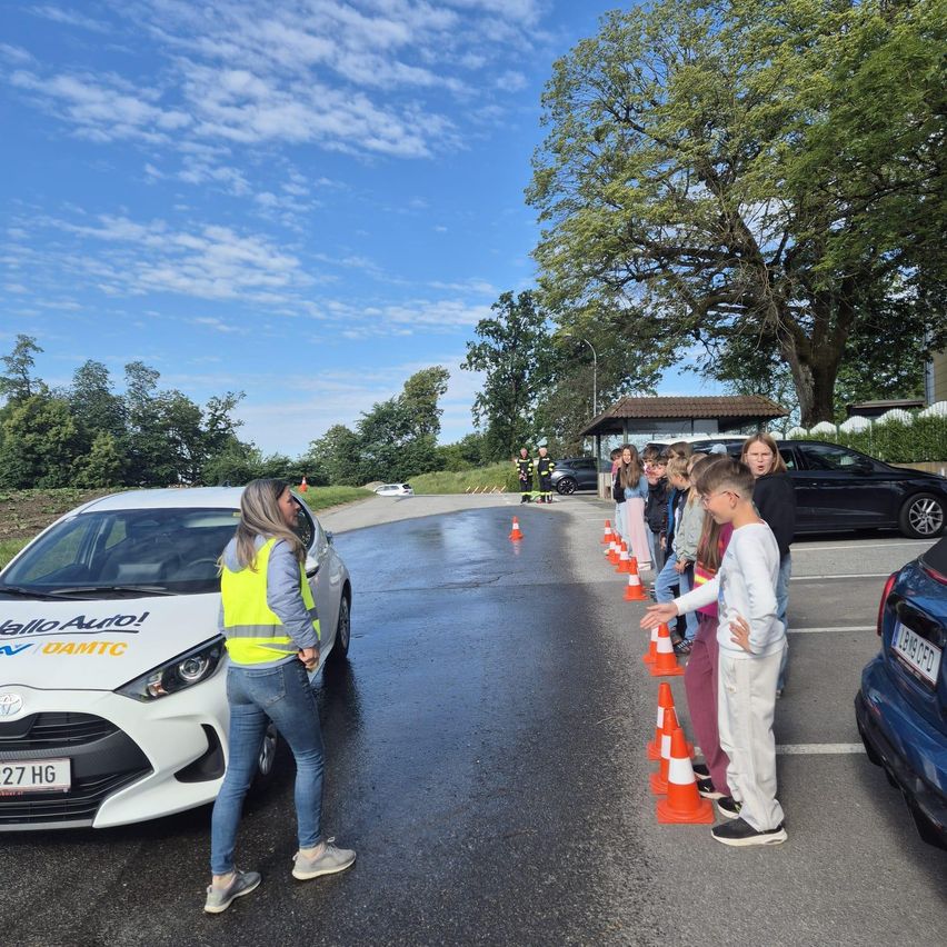 Eine Frau in einer Warnweste steht neben einem weißen Toyota. Eine Gruppe von Kindern steht in einer Reihe vor Verkehrskegeln. Ein Feuerwehrauto ist in der Nähe geparkt.