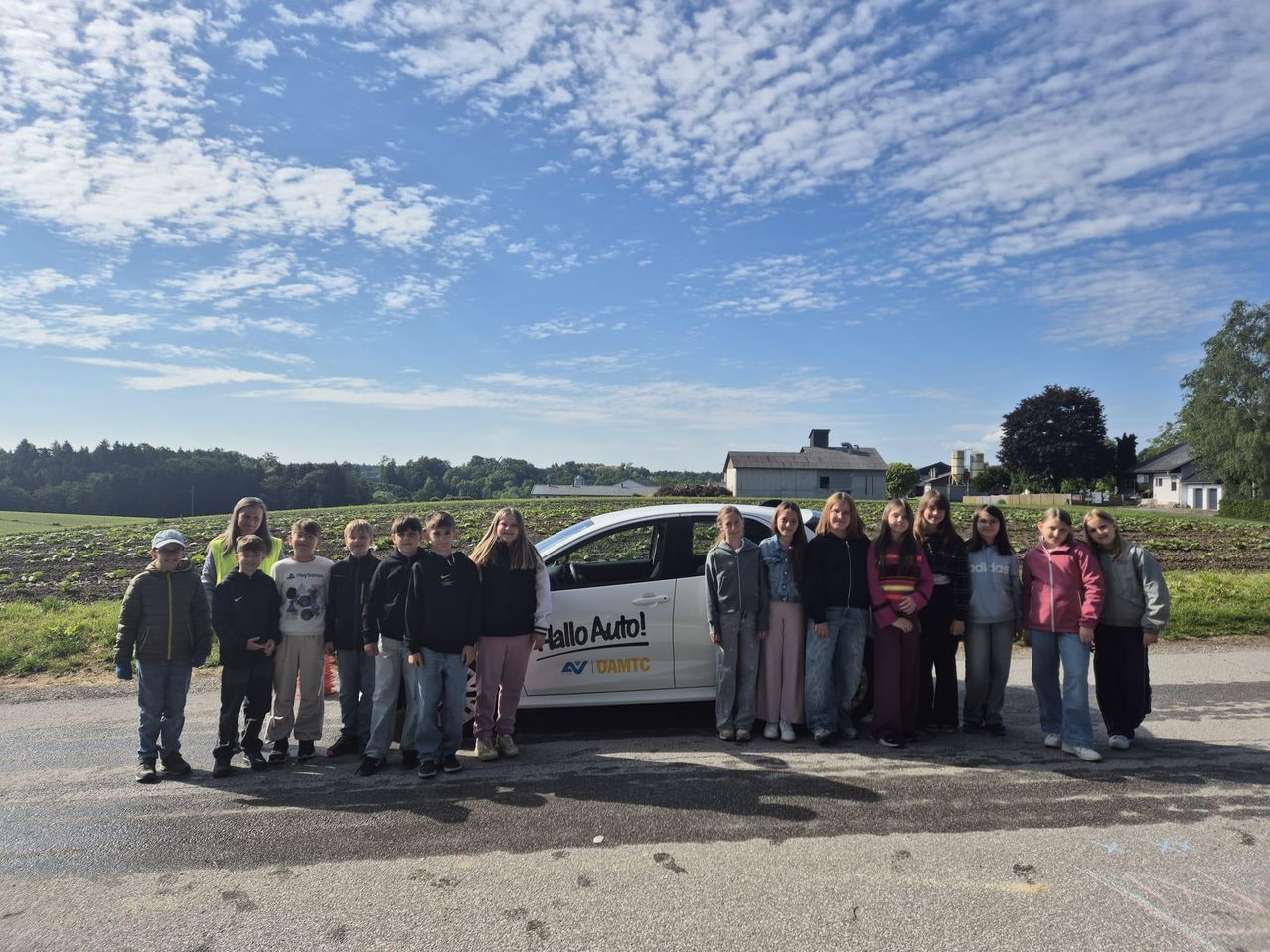 Eine Gruppe junger Personen, sowohl Jungen als auch Mädchen, stehen vor einem weißen Fahrzeug mit dem Text Hallo Auto! auf dem Land.