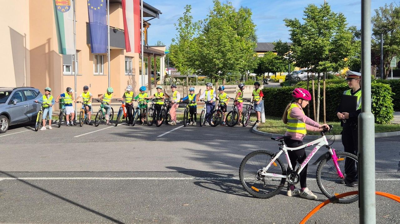 Eine Gruppe von Kindern in gelben Westen und Helmen fährt mit Fahrrädern auf einer Straße, wobei ein Mädchen vorne einen rosa Helm trägt. Bäume und Gebäude säumen die Straße.