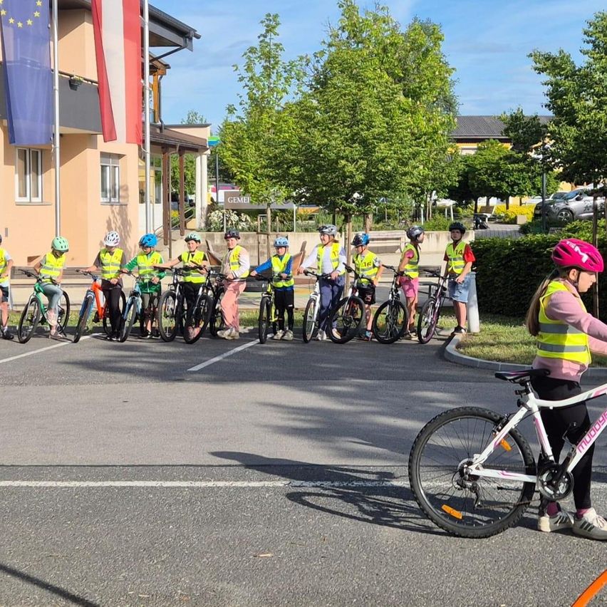 Eine Gruppe von Kindern in gelben Westen und Helmen fährt mit Fahrrädern auf einer Straße, wobei ein Mädchen vorne einen rosa Helm trägt. Bäume und Gebäude säumen die Straße.