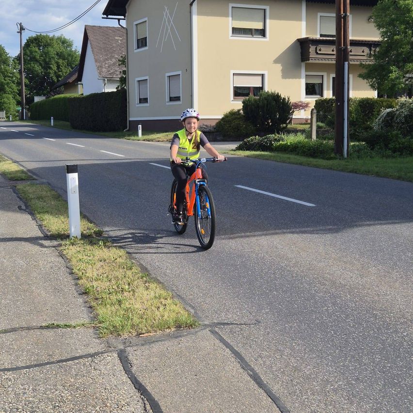 Ein junges Kind fährt mit einem Fahrrad eine Wohnstraße hinunter, mit Häusern und Grünflächen im Hintergrund.
