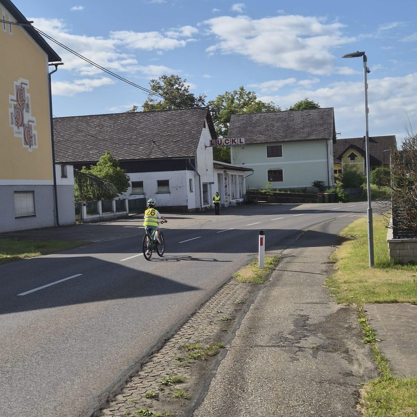 Ein Radfahrer in einem gelben Weste fährt eine leere Straße entlang. Gebäude säumen die Straße, und eine Person in einer reflektierenden Weste steht in der Nähe eines Schildes. Der Himmel ist bewölkt.