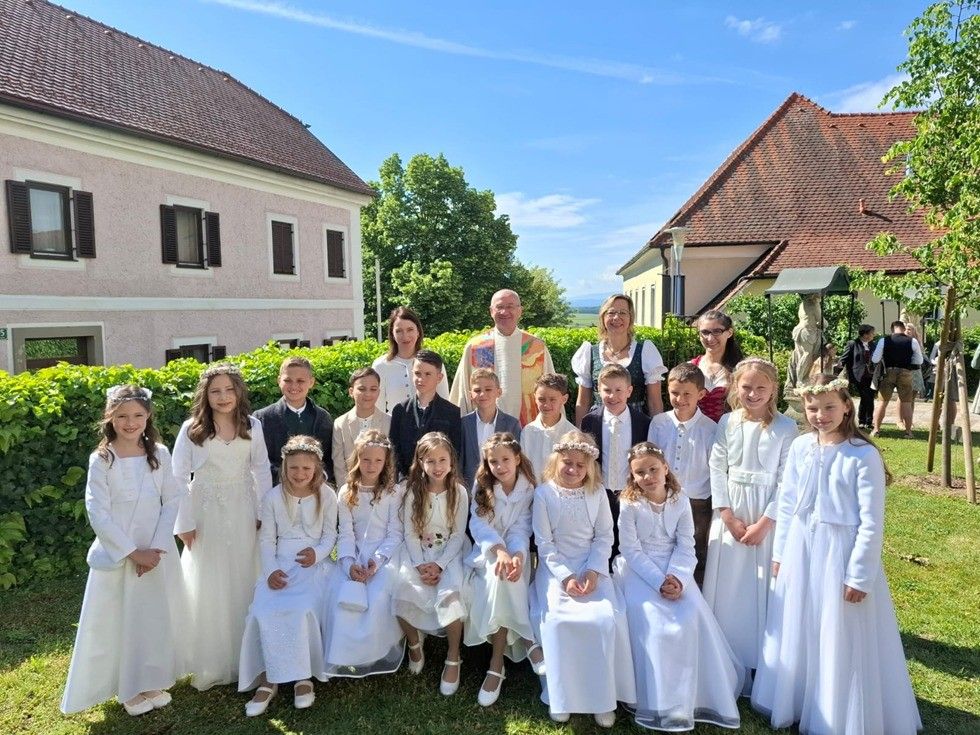 Eine Gruppe von Kindern in weißen Kleidern und Blumenkränzen posiert für ein Foto in einem Garten mit zwei Erwachsenen und Gebäuden im Hintergrund.