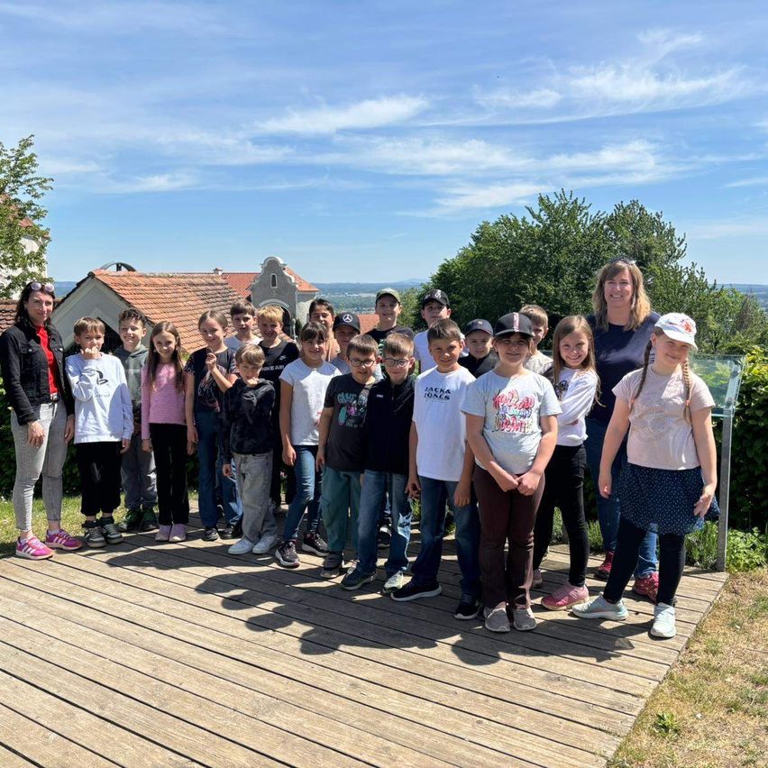 Eine Gruppe von Kindern und Erwachsenen, darunter zwei Frauen, posiert für ein Foto auf einer Holzterrasse mit einer malerischen Kulisse.