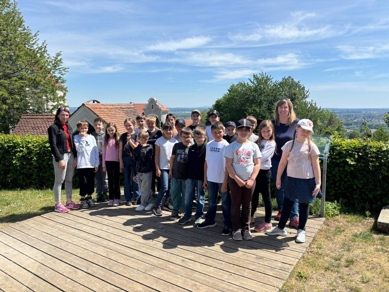 Eine Gruppe von Kindern und Erwachsenen, darunter zwei Frauen, posiert für ein Foto auf einer Holzterrasse mit einer malerischen Kulisse.