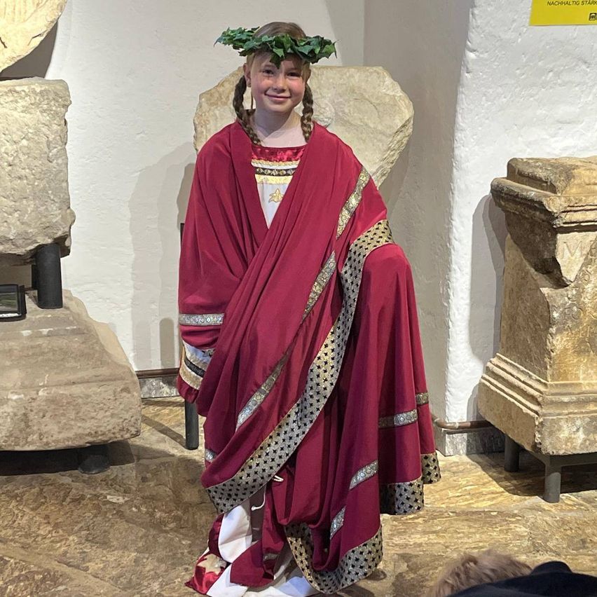 Ein junges Mädchen mit geflochtenen Haaren trägt ein rotes und goldenes römisches Kostüm. Sie steht zwischen Steinskulpturen und einer Wand mit einem gelben Schild.