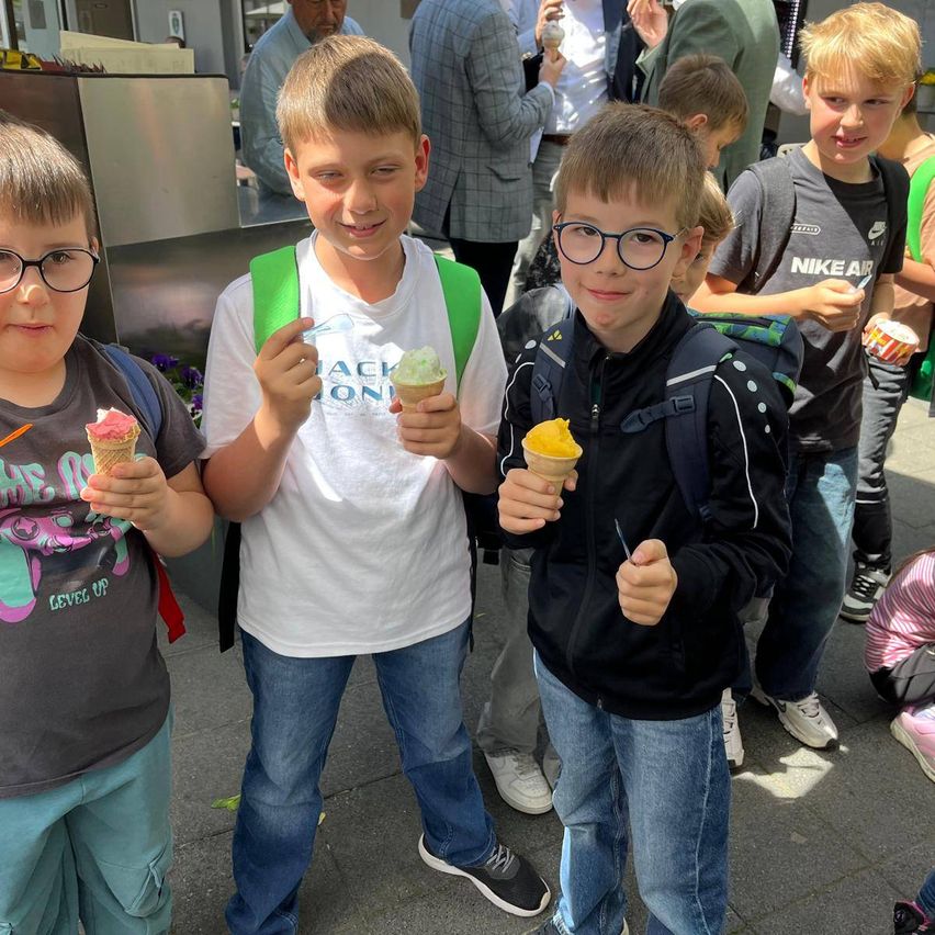 Drei Jungen in Freizeitkleidung halten Eisbecher in der Hand und stehen draußen, mit weiteren Kindern und Erwachsenen dahinter.