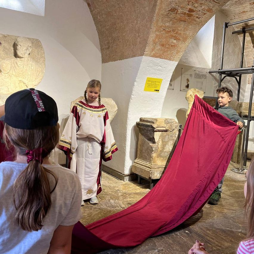 Eine Gruppe von Kindern ist in einem Museum mit einer Steinstatue im Hintergrund. Ein Kind trägt ein Kostüm, während ein anderes ein rotes Tuch hält.