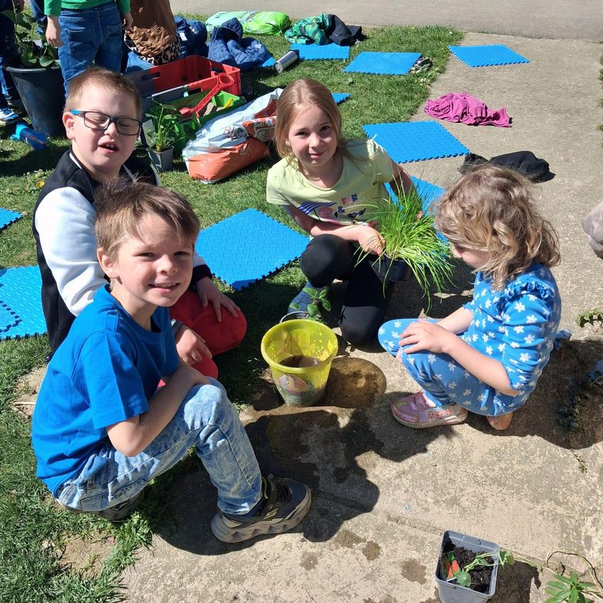 Vier Kinder pflanzen Gras in einem Garten. Die Kinder sitzen auf dem Boden und halten einen Eimer mit Wasser. Eines von ihnen trägt eine Brille.