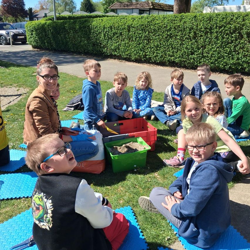 Eine Gruppe von Kindern und einer erwachsenen Frau sitzt auf einem Rasen, einige mit Sandsandkasten, in der Nähe eines Hecken und einer Straße.