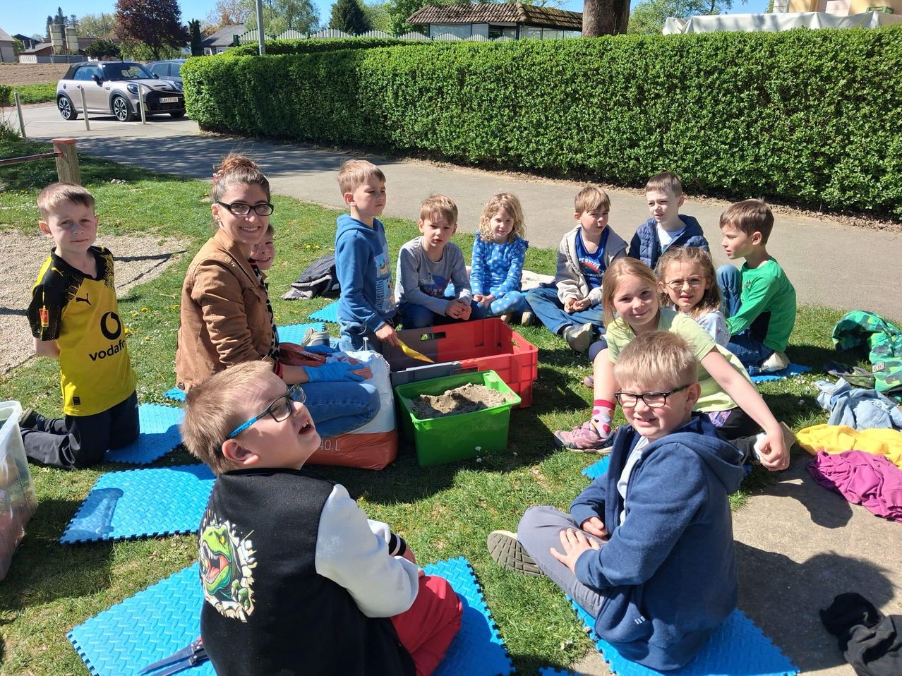 Eine Gruppe von Kindern und einer erwachsenen Frau sitzt auf einem Rasen, einige mit Sandsandkasten, in der Nähe eines Hecken und einer Straße.