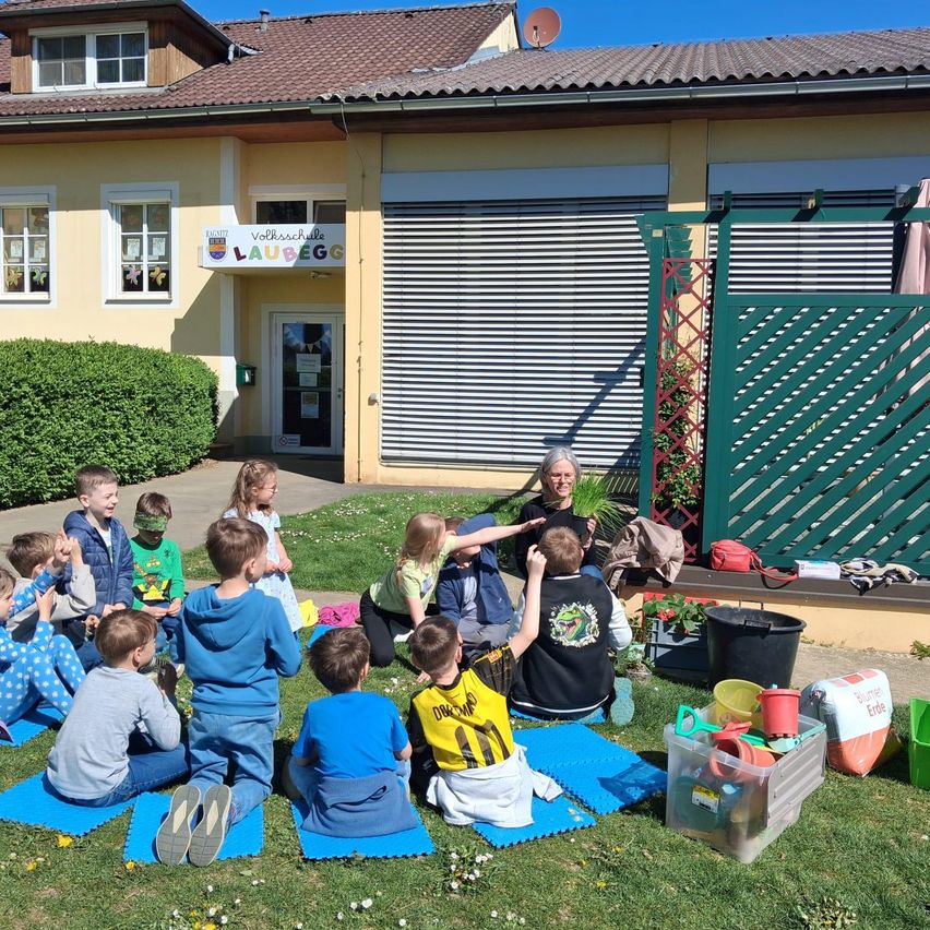 Eine Gruppe von Kindern sitzt kreisförmig auf dem Rasen vor einem Gebäude. Eine erwachsene Frau steht hinter ihnen, möglicherweise im Gespräch.