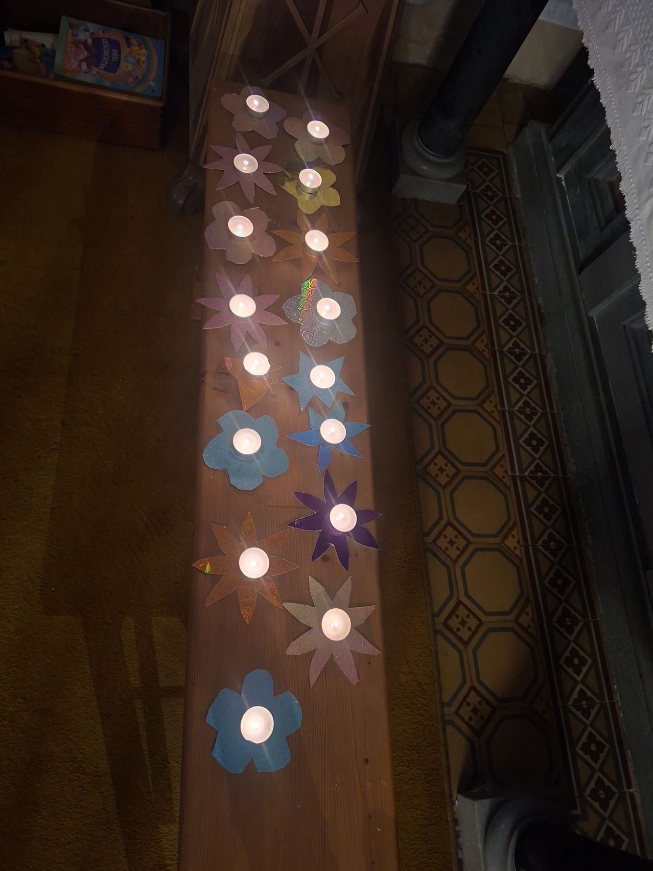 Eine Holzbank ist mit mehrfarbigen Papierblumen verziert, jede mit einer brennenden Kerze, in einer Reihe auf einem Teppichboden mit einem gefliesten Muster angeordnet.