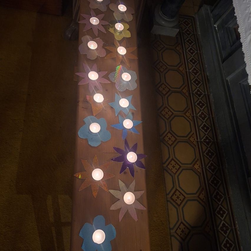 Eine Holzbank ist mit mehrfarbigen Papierblumen verziert, jede mit einer brennenden Kerze, in einer Reihe auf einem Teppichboden mit einem gefliesten Muster angeordnet.