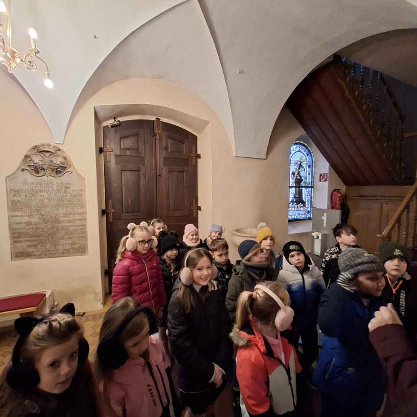Eine Gruppe von Kindern, die Winterjacken und Ohrenschützer tragen, steht in einer alten Kapelle. Hinter ihnen befindet sich eine Holztür, und rechts ist eine Treppe mit einem Feuerlöscher. Eine Tafel mit Text steht an der Wand in der Nähe der Tür.