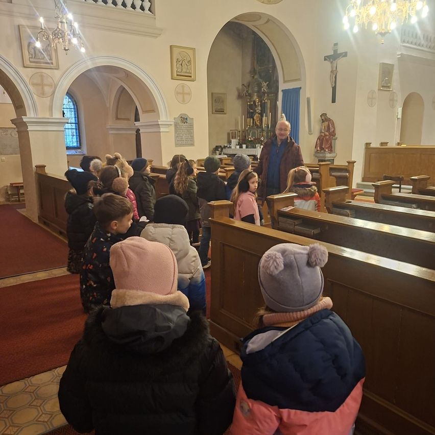 Eine Gruppe von Kindern und ein Mann sind in einer Kirche und schauen zu einem Mann, der spricht. Die Kirche hat Holzbänke, einen roten Teppich und religiöse Dekorationen.