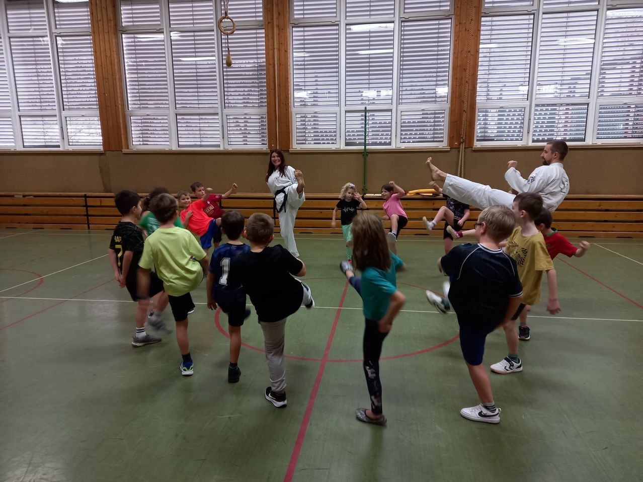 In einer Turnhalle demonstriert eine Frau in einer Karate-Ausrüstung einen Tritt, während Kinder sie nachahmen, einige tragen nummerierte Trikots. Die Turnhalle hat Holzwände, Fenster und einen Ring, der von der Decke hängt.