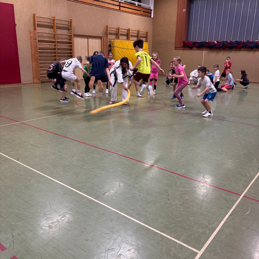In einer Turnhalle spielen mehrere Kinder ein Spiel. Sie rennen, einige halten einen gelben Stock, und andere jagen. Die Halle hat einen weißen Boden mit Markierungen.