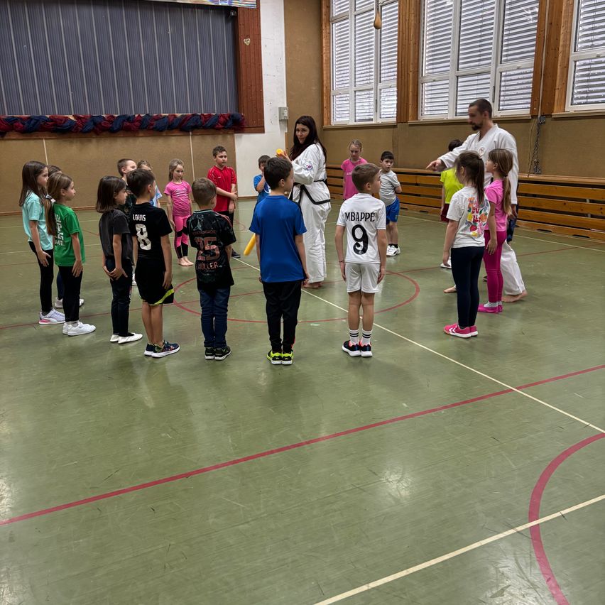 Eine Gruppe junger Kinder versammelt sich mit einem Mann und einer Frau in einer Turnhalle. Sie tragen Turnschuhe und einige haben Nummern auf ihren Trikots.