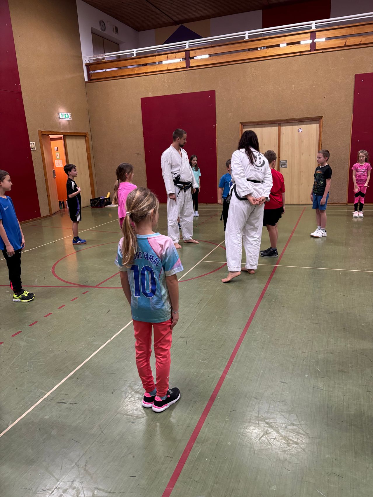 Ein Karate-Kurs mit Kindern und zwei Ausbildern in einer Turnhalle. Die Kinder tragen nummerierte Shirts und Turnschuhe und stehen im Kreis. Die Ausbilder sind in weißen Uniformen.
