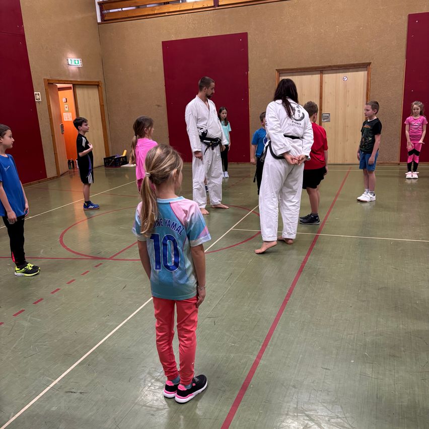 Ein Karate-Kurs mit Kindern und zwei Ausbildern in einer Turnhalle. Die Kinder tragen nummerierte Shirts und Turnschuhe und stehen im Kreis. Die Ausbilder sind in weißen Uniformen.
