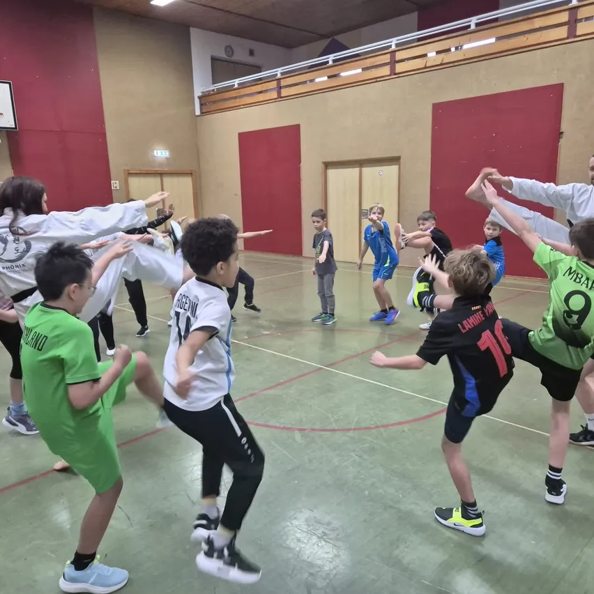 In einer Indoor-Sporthalle üben Kinder in Sportuniformen verschiedene Übungen. Ein Junge in einem grünen Trikot streckt seine Arme aus. In der Nähe sind Kinder in weißen und blauen Trikots bei einer Aktivität.