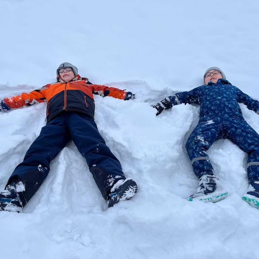 Zwei Kinder liegen im Schnee, eines in Orange und Schwarz, das andere in Blau, beide tragen Winterkleidung und Brillen.