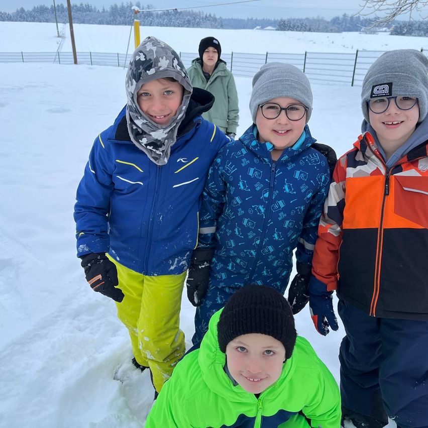 Fünf Kinder stehen lächelnd im Schnee. Drei von ihnen tragen blaue Jacken, eines eine grüne Jacke und eines eine orange-schwarze Jacke. Sie sind draußen in einem schneebedeckten Feld.