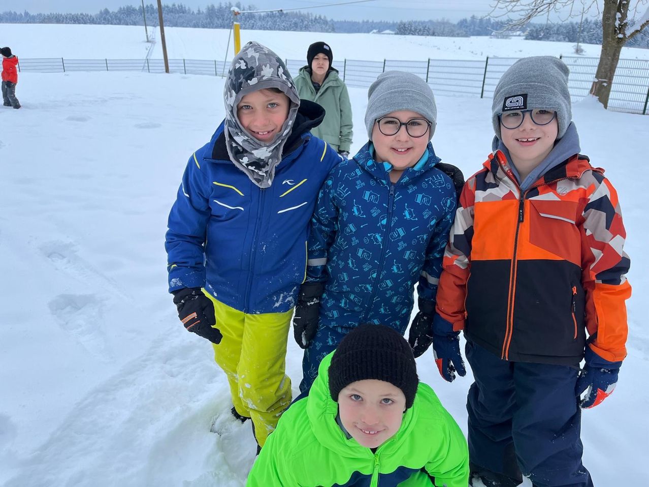 Fünf Kinder stehen lächelnd im Schnee. Drei von ihnen tragen blaue Jacken, eines eine grüne Jacke und eines eine orange-schwarze Jacke. Sie sind draußen in einem schneebedeckten Feld.