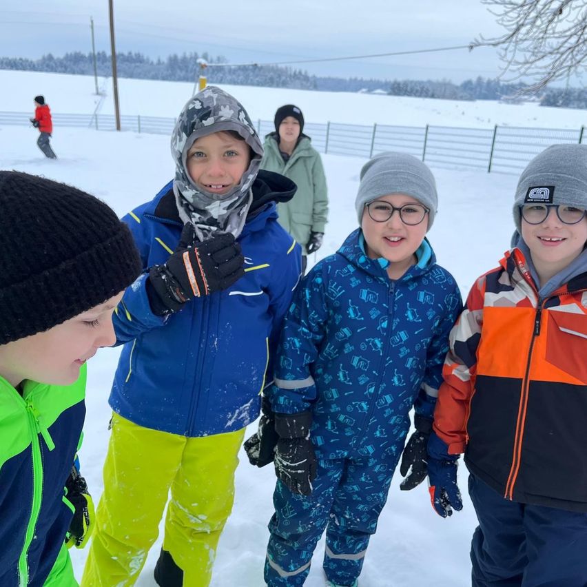 Vier Kinder stehen im Schnee und tragen Winterkleidung. Eines trägt eine blaue Jacke und gelbe Hose, ein anderes ein blaues Overall mit Brille. Zwei andere tragen schwarze Mützen. Im Hintergrund skizziert eine Person.